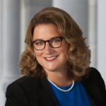 Headshot of Heather smiling directly at the camera wearing a black suit coat on a gray pillar backdrop.