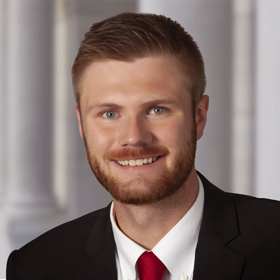 Headshot of Jim smiling directly at the camera wearing a black suit and a red tie on a gray backdrop.