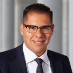 Headshot of Gil Gonzalez smiling directly at the camera. He is wearing a navy suit coat with a white dress shirt and a blue tie.