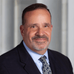Professional headshot of Ron Menna in a dark suit jacket, light blue dress shirt, and patterned tie, posed against a softly blurred indoor background with light-colored walls.