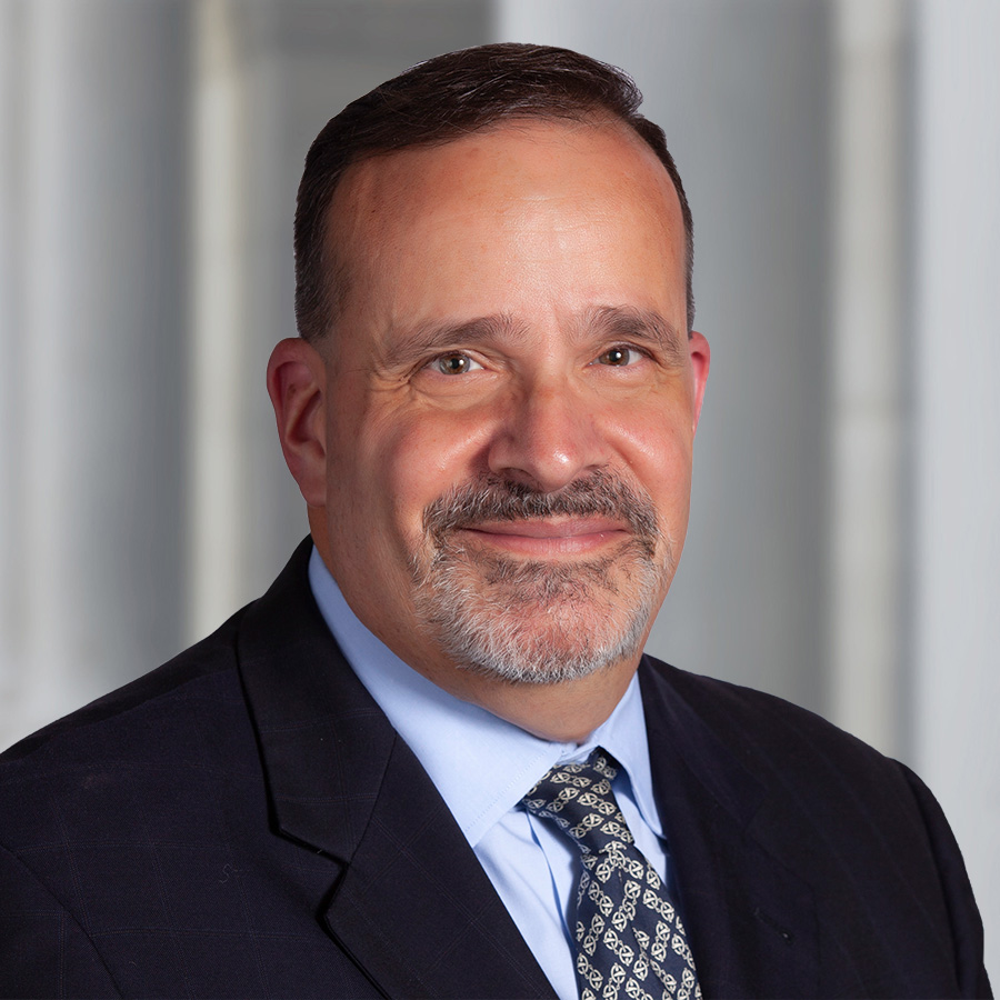 Professional headshot of Ron Menna in a dark suit jacket, light blue dress shirt, and patterned tie, posed against a softly blurred indoor background with light-colored walls.