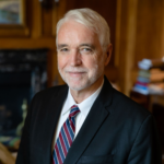Professional headshot of Timothy Killeen wearing a dark suit jacket, white collared shirt, and striped tie, posed in front of a softly blurred indoor background with wood paneling and shelves.
